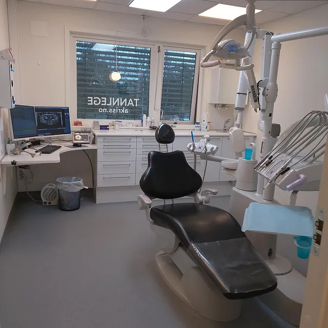 Tannlegekontor med svart behandlingsstol, monitor, instrumenter og datamaskin ved vindu.